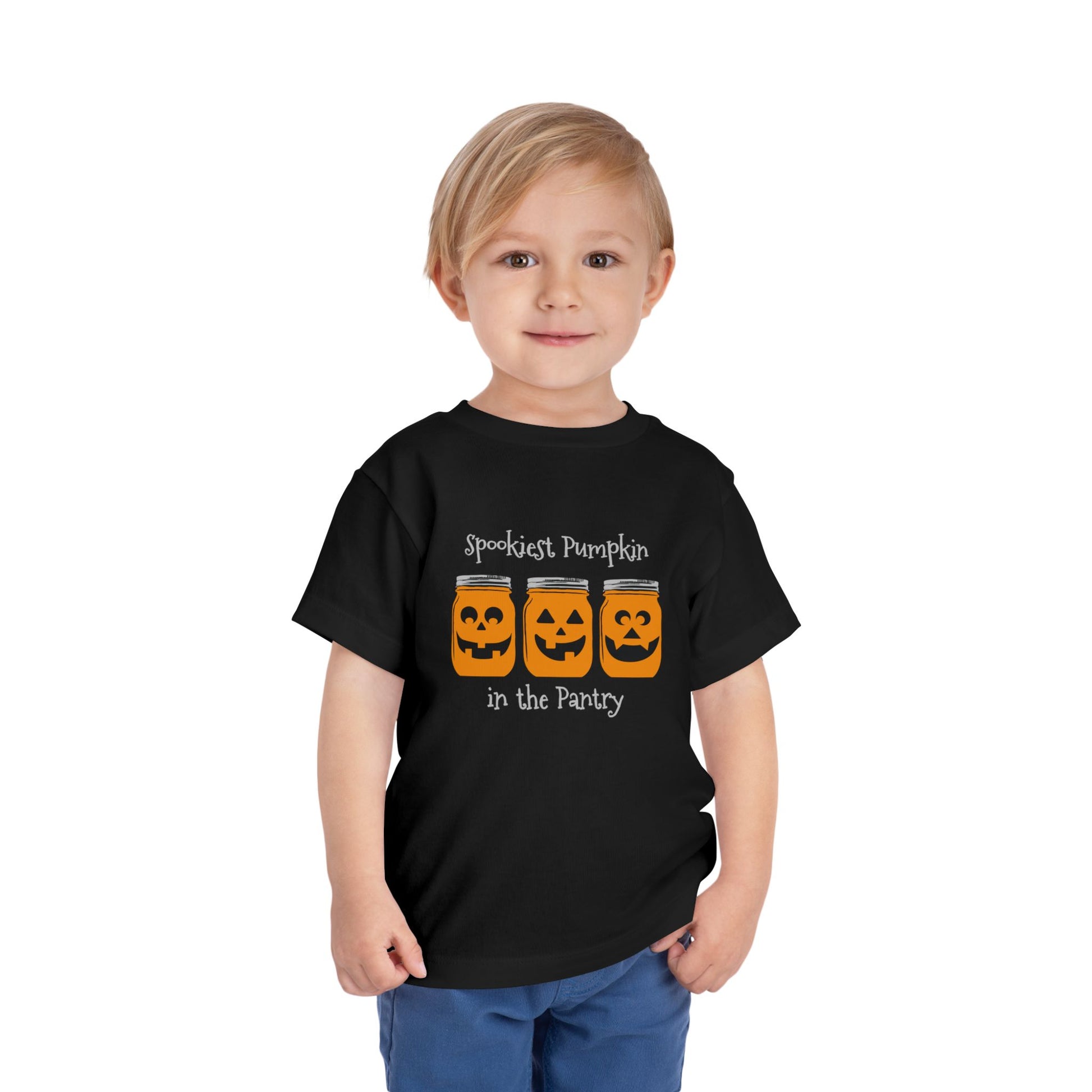 A young child beams in the Printify Toddler Tee Halloween Spookies Pumpkin in the Pantry—featuring three jack-o'-lantern jars and festive text, it’s the perfect spooky outfit for your little one this Halloween!.