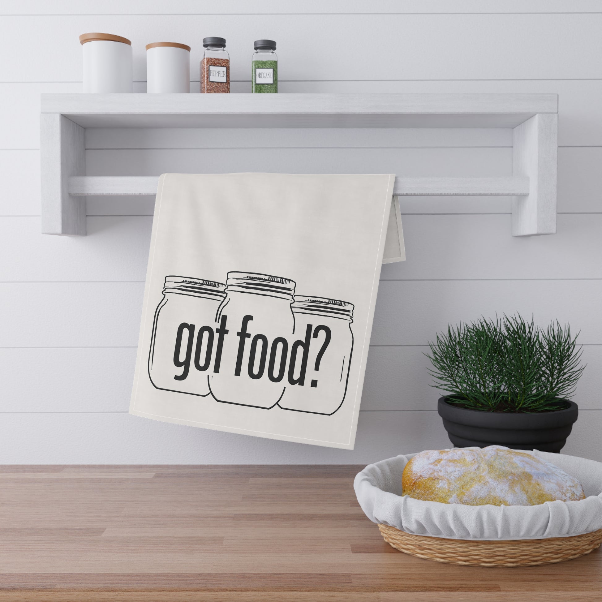 The Printify Got Food? Kitchen Tea Towel, featuring three mason jars and "got food?", hangs above a wooden counter, bringing playful charm to your kitchen decor next to a potted plant and a towel-covered bowl.