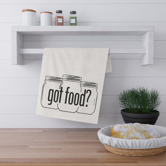 The Printify Got Food? Kitchen Tea Towel, featuring three mason jars and "got food?", hangs above a wooden counter, bringing playful charm to your kitchen decor next to a potted plant and a towel-covered bowl.