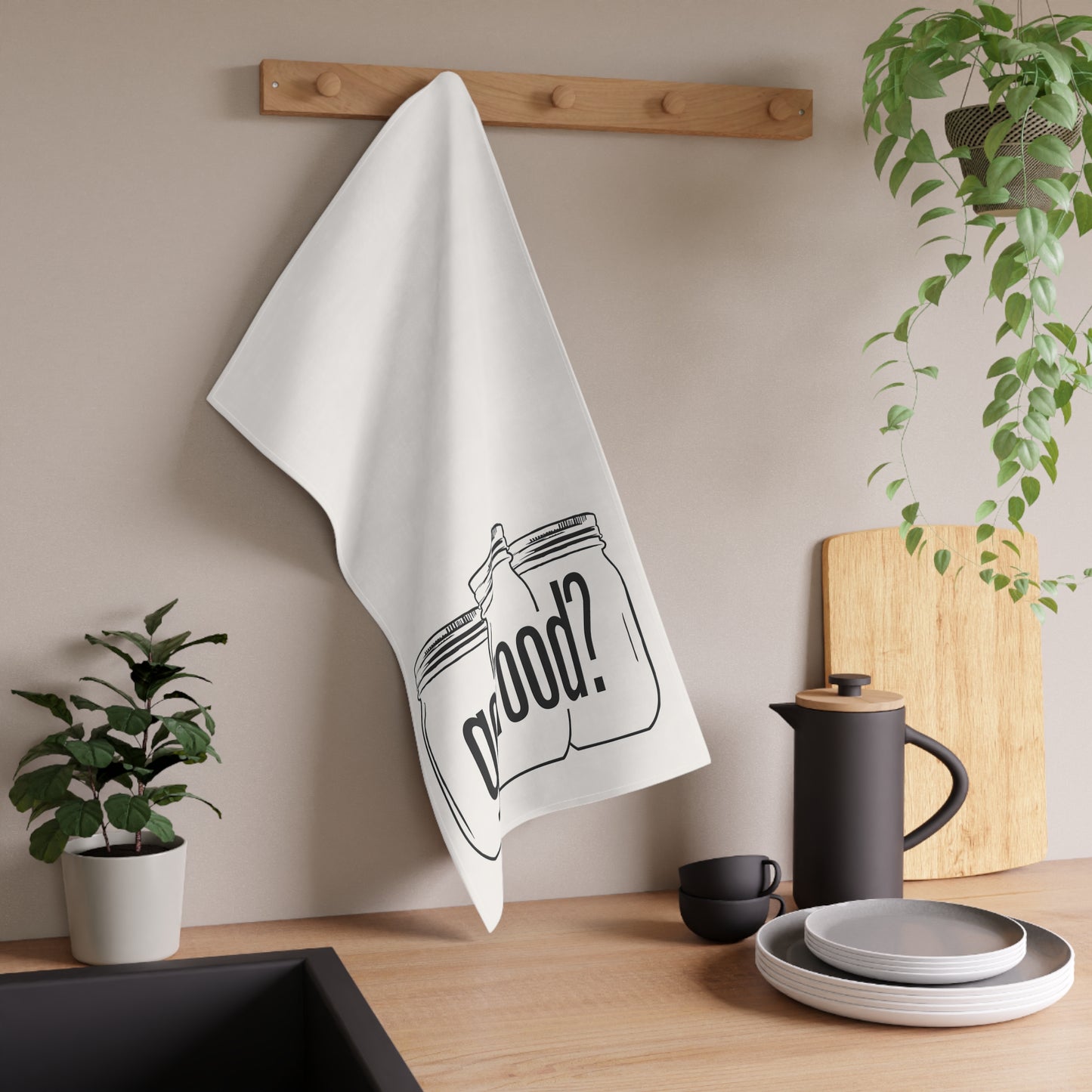 The Got Food? Kitchen Tea Towel by Printify hangs from a wooden peg rack above a sink, adding quirky decor to the kitchen alongside plants, a coffee pot, cups, and plates on the counter.