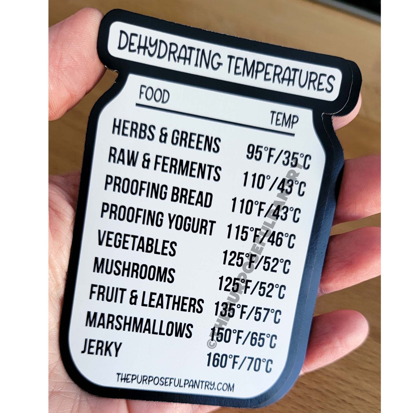 A person is holding a label that says The Purposeful Pantry Dehydrator Temperature Guide Refrigerator Magnet.